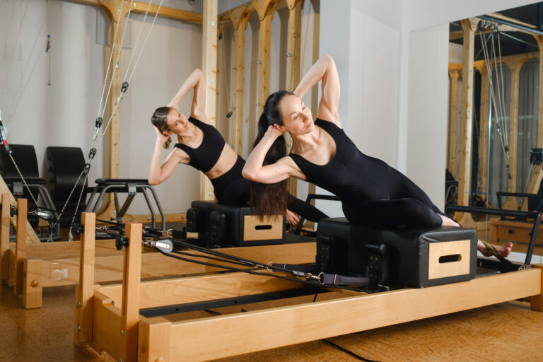 Two women balance on a pilates reformer machine in the gym